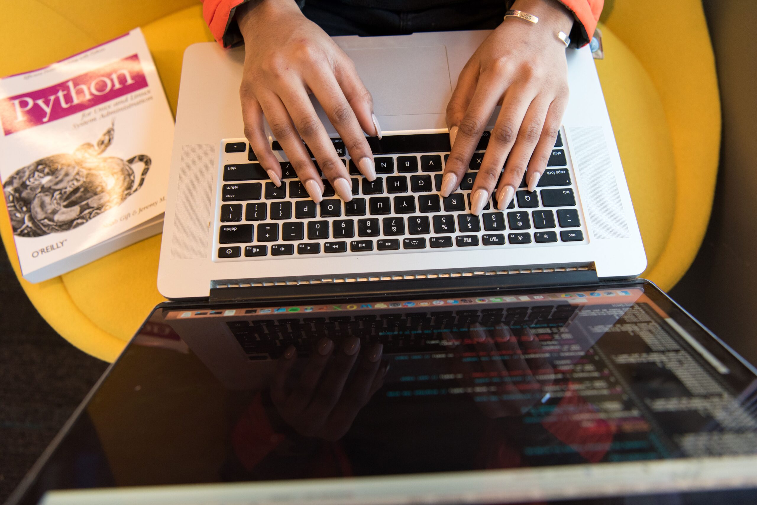 Understanding the Python Developer Role: Responsibilities, Skills & Career Opportunities Close-up of hands typing on a laptop keyboard, Python book in sight, coding in progress.