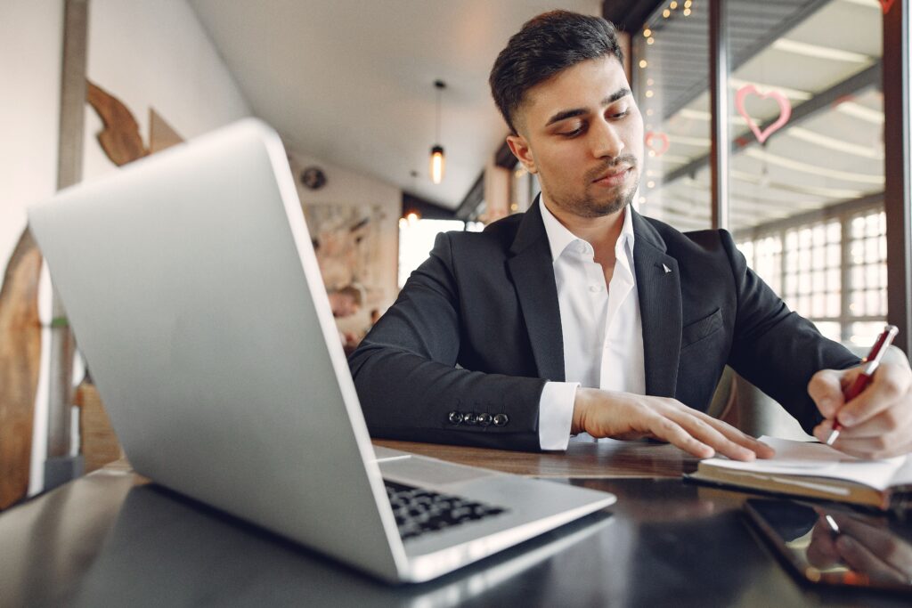 From below of concentrated ethnic male in formal wear sitting in creative workplace and taking notes while using laptop and working on business project