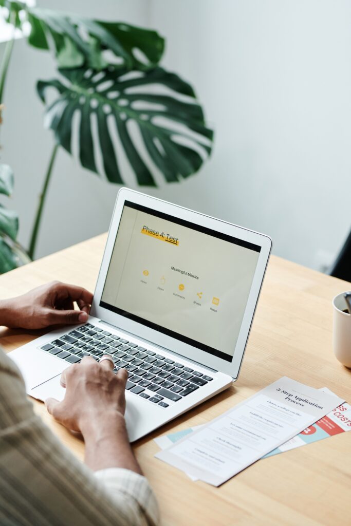 Adult using a laptop on a wooden desk with a large plant leaf in the background, creating a serene workspace.